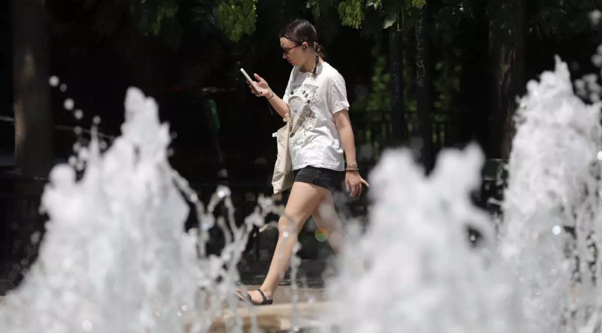 Tânără surprinsă în timp ce trece pe lângă o fântână arteziană. Se uită în telefon și are căști în urechi. Poartă un tricou alb, o pereche de pantaloni scurți negri și sandale.