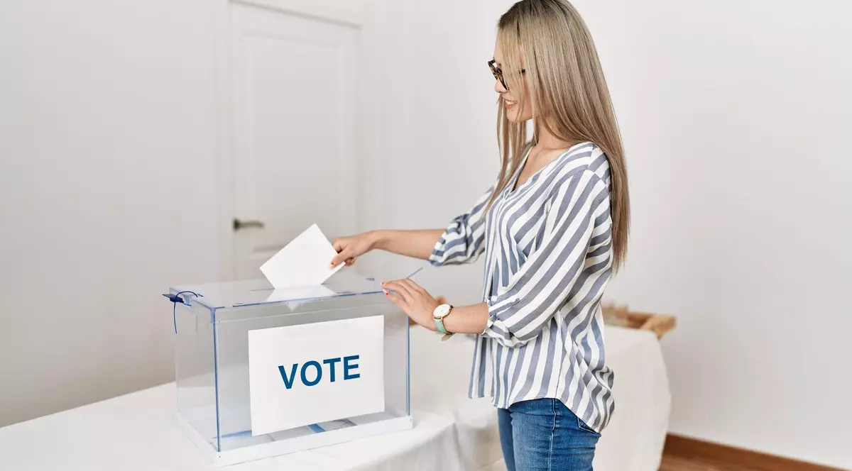 Tânără blondă, care poartă o cămașă albă cu dungi albastre și blugi albaștri, surprinsă în timp ce introduce buletinul de vot într-o urnă