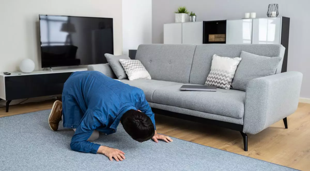 Un bărbat intr-o cîmașă albastra sta in genunchi in casă, pe un covor gri, si se uită sub canapeaua tot cu, cu pernute decorative albe, cautând ceva pierdur, in spate se vede un televizor si un dulap alb