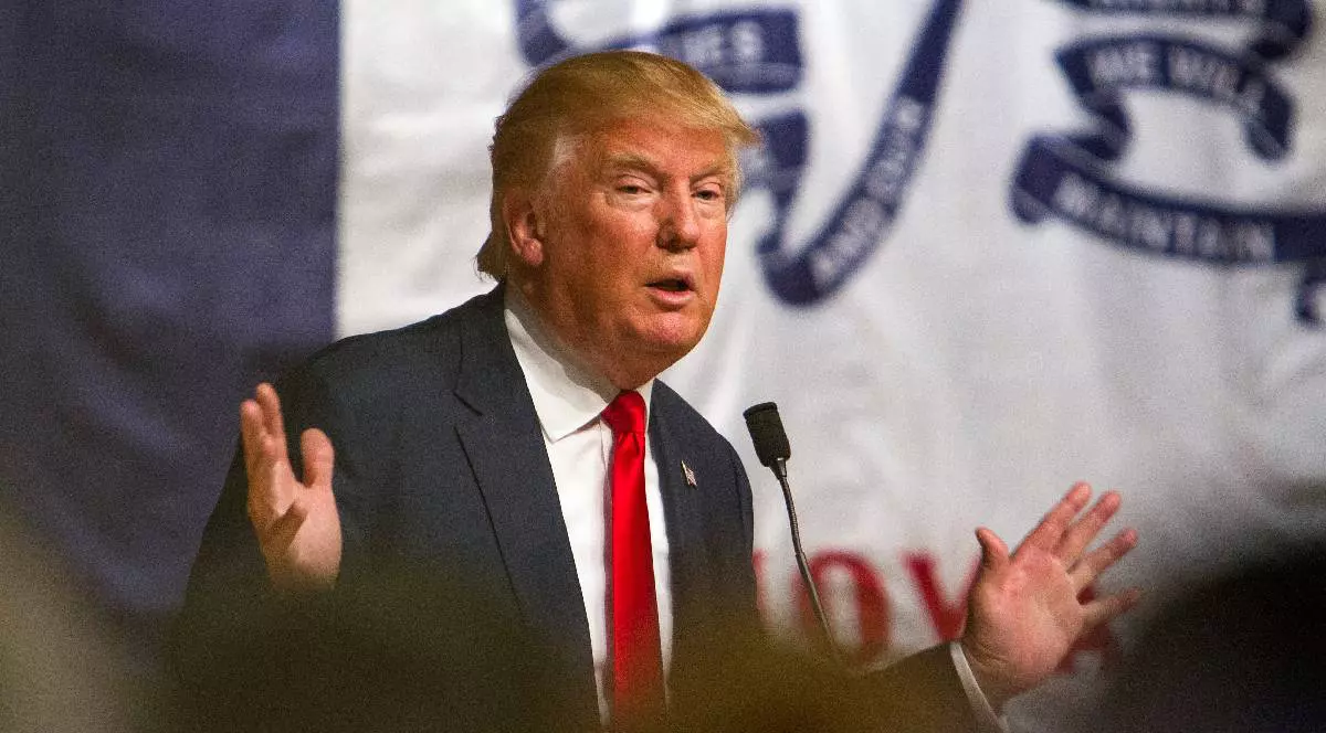 În imagine, Donald Trump ține mâinile larg deschise în timp ce explică ceva la un microfon.