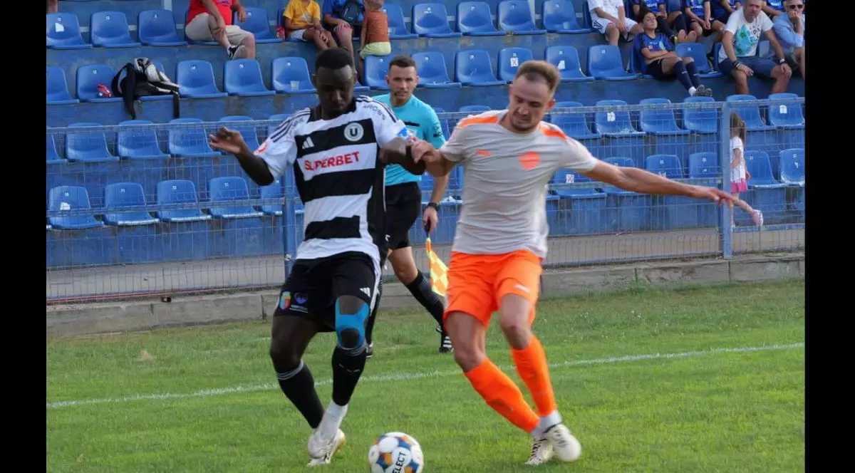 doi jucători se luptă pentru controlul mingii, care se află pe gazon, între ei. Unul dintre jucători poartă un echipament cu tricou alb cu dungi negre orizontale și pantaloni scurți negri, iar celălalt este îmbrăcat într-un tricou deschis la culoare și pantaloni scurți portocalii.