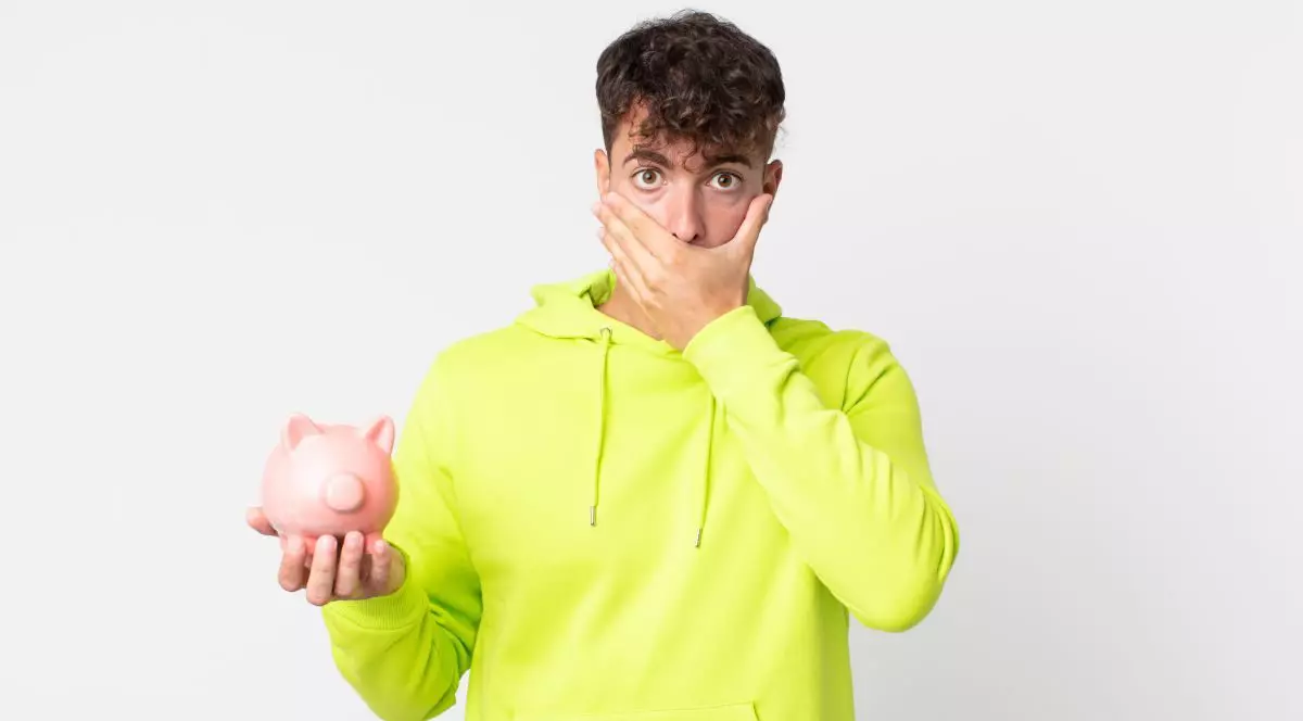 Un tânăr îmbrăcat cu hanorac verzui își acoperă partea de jos a feței cu mâna, fiind speriat sau șocat, iar în cealaltă mână ține o pușculiță roz în formă de porc