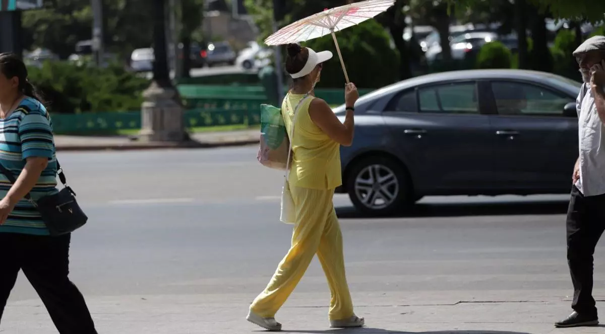 În centrul imaginii este o femeie îmbrăcată în galben, cu o șapcă albă și umbrelă de soare deasupra capului, în dreapta este un bărbat care vorbește la telefon, iar în stânga o femeie. În spate, se vede o mașină pe stradă, București, 28 iunie 2025