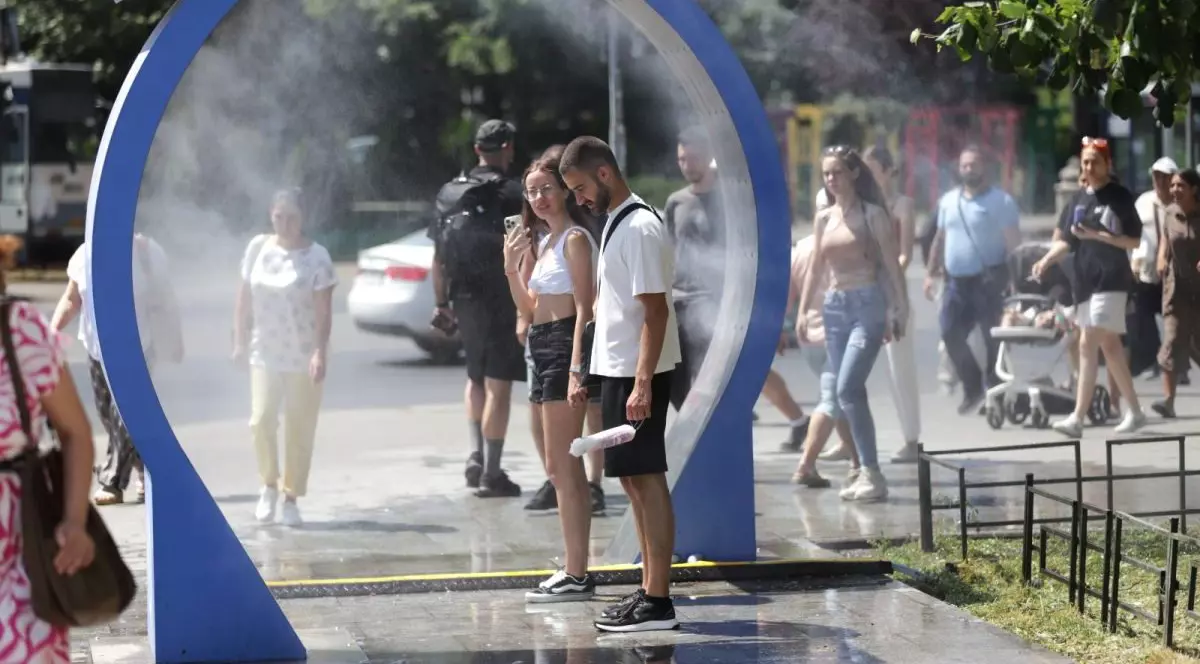 Doi tineri stau în interiorul unui duș stradal, iar alți oameni trec prin acea zonă, într-o zi caniculară de vară, București, 28 iunie 2025