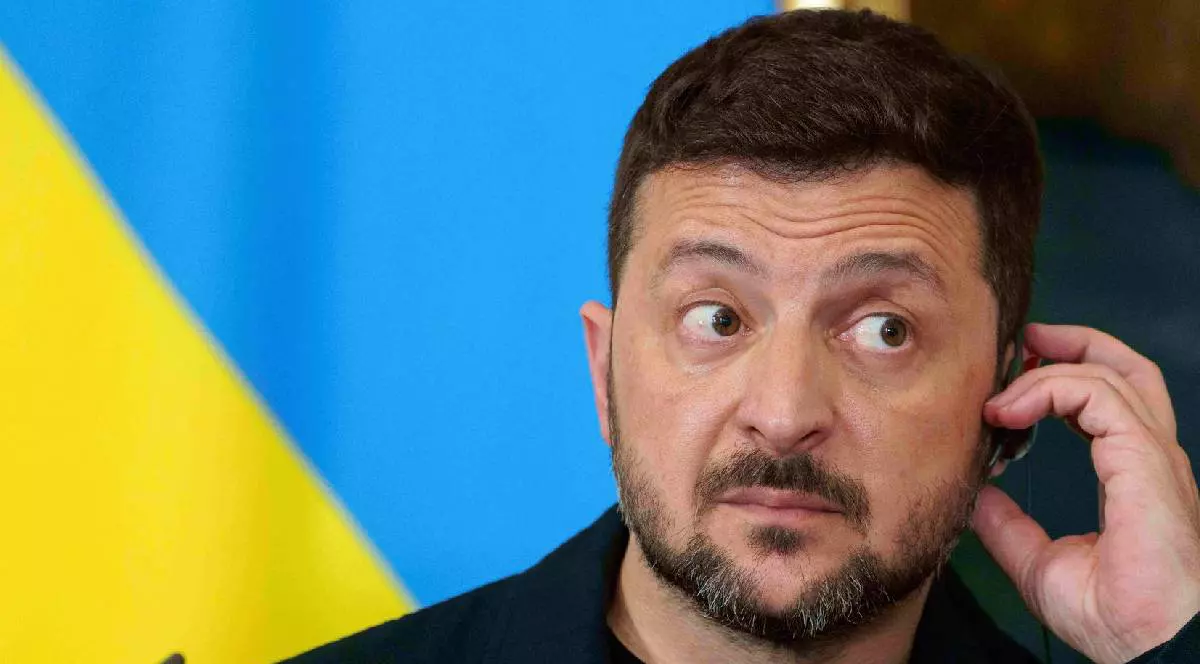 În imagine, Volodimir Zelenski ascultă ceva într-o cască și pare nedumerit. Pe fundal e steagul Ucrainei