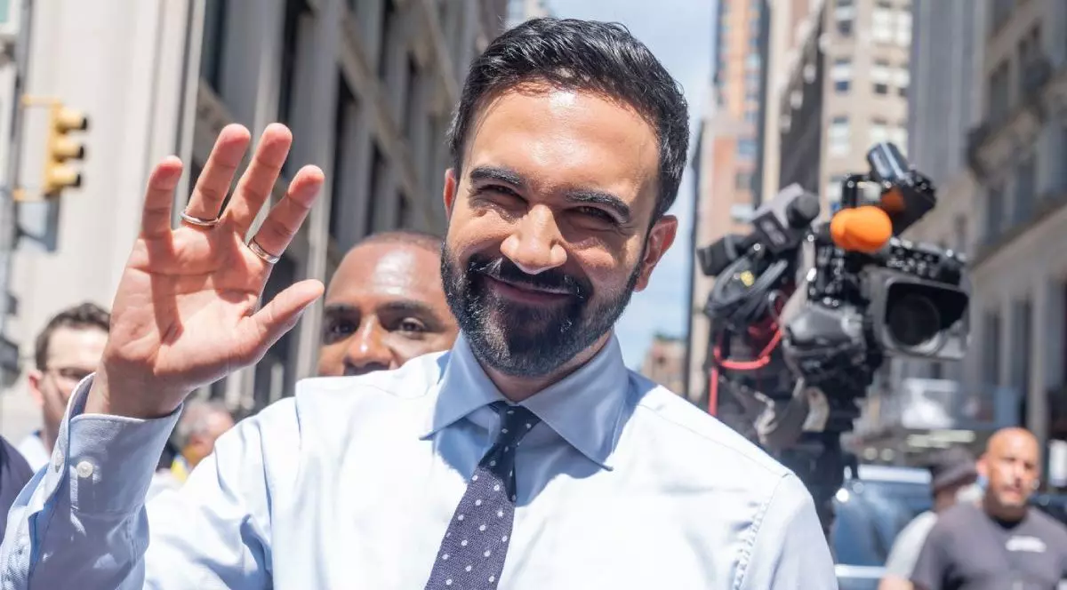 Zohran Mamdani, [n în cămasă alba, cravata gri cu picateșe, cu mana dreapta ridicata in semn de salut, participă la parada Pride din New York, 29 iunie 2025, in spatele lui se vad camere video si jurnalisti