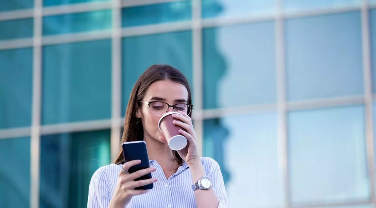 Femeie tanara care isi bea cafeua si se uita pe telefon. Se afla in fata unei cladiri de birouri