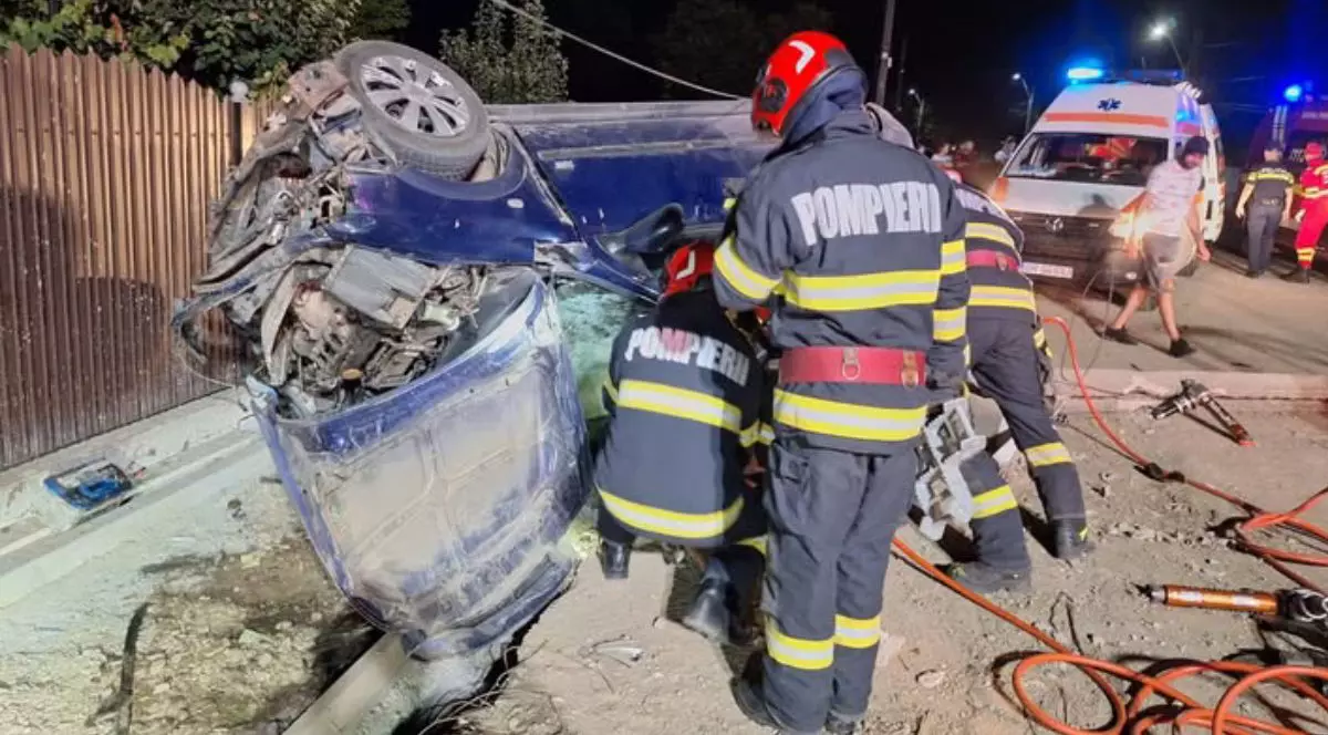 In fotografia de noapte se vad doi pompieri, unul in picioare, unul pe vine, langă o masină distrusa intr0un accident, in spare este o ambulanță, apamedici si oameni