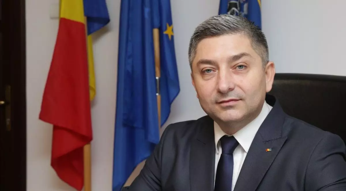 Alin Tișe, îmbrăcat într-un costum gri, în timp ce stă la birou și are pe fundal steagurile României și Uniunii Europene