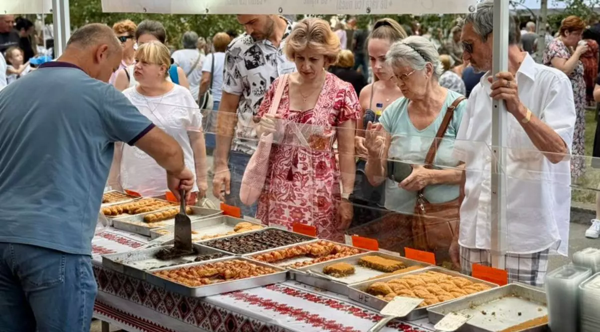 În imagine sunt vizibile mai multe tăvi metalice așezate pe o masă lungă, fiecare tavă conținând diferite tipuri de prăjituri sau produse de patiserie turcești. În partea stângă, un bărbat poartă un tricou bleu și ține o spatulă metalică deasupra unei tăvi. De cealaltă parte a mesei, câteva persoane stau grupate în așteptare, unele ținând genți sau sacoșe, spatele lor fiind acoperit parțial de o bară transparentă de protecție