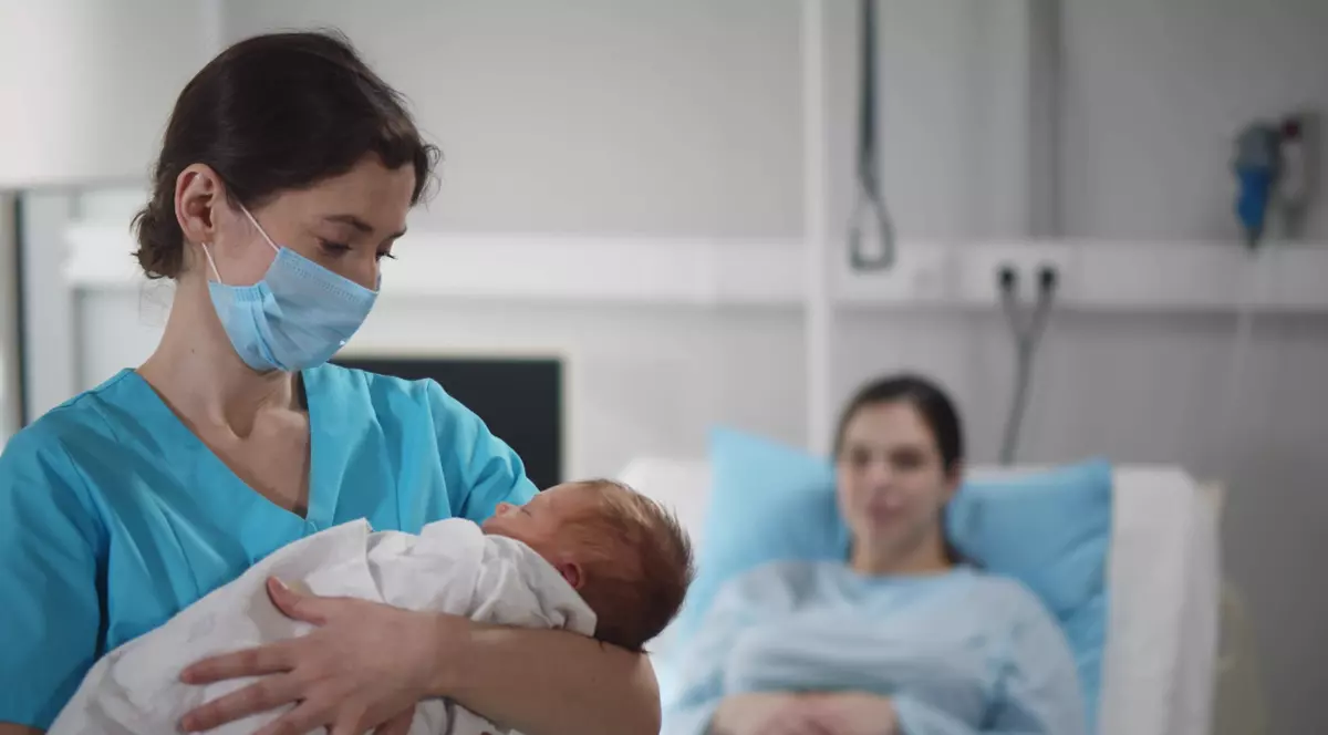 Portret al unei asistente medicale purtând mască de protecție, ținând în brațe un nou-născut, alături de mama acestuia, care se odihnește în patul de spital