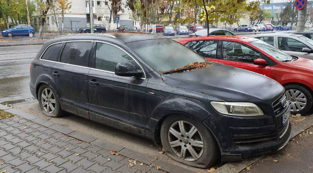 Audi Q7, de culoare neagră, cu roțile dezumflate, abandonat pe stradă, lângă alte mașini