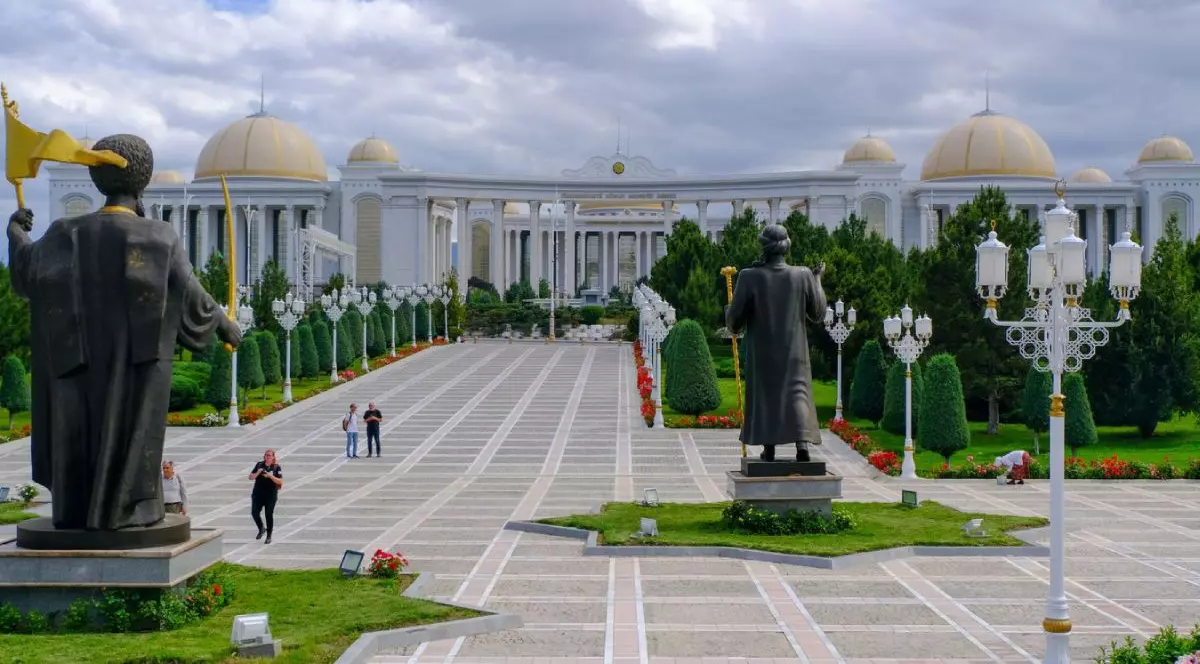 Biblioteca Națională din Așgabat, capitala Turkmenistanului Se vede o cladire impozanta, cu multe corpuri, alb[ cu acoperisuri aurite, alei largi pe care se plimba oameni, marginite de zone verzi, si doua statui care se vad din spate.