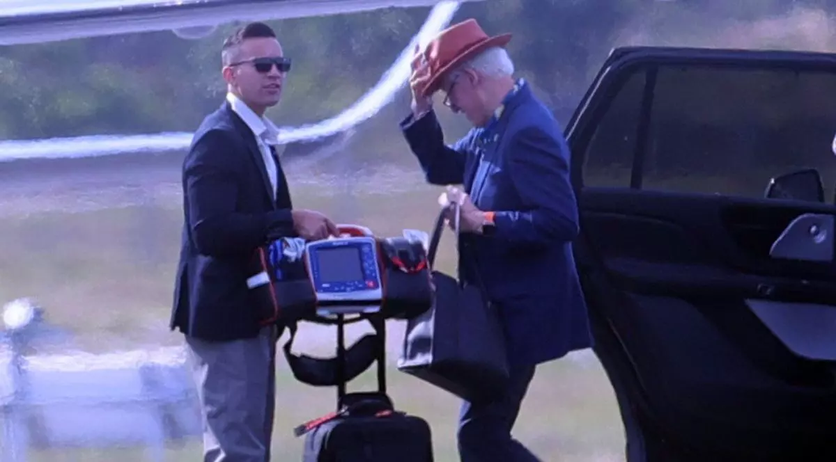 Bill Clinton, cu mâna pe pălărie, însoțit de un reprezentant al personalului său de securitate, este pe un aeroport și are o geantă cu un defibrilator portabil, Hamptons, New York, Statele Unite, 28 august 2025