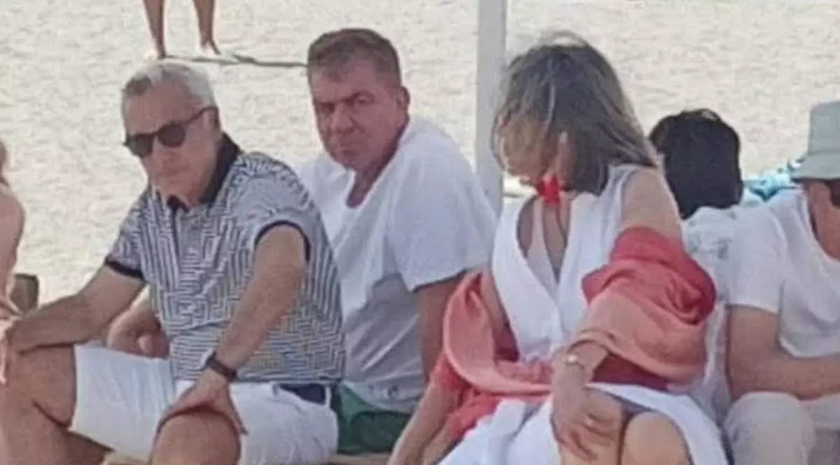 Călin Georgescu (stânga) și Cristela Georgescu (dreapta) surprinși în timp ce stau pe șezlong la mare. Fostul candidat la prezidențiale poartă ochelari de soare, un tricou și pantaloni albi.