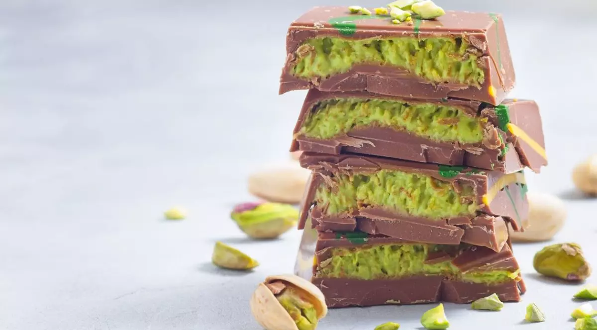 Batoane de ciocolată cu lapte cu cremă de fistic și umplutură kadaifi, așezate pe o masă cu fistic întreg și tocat