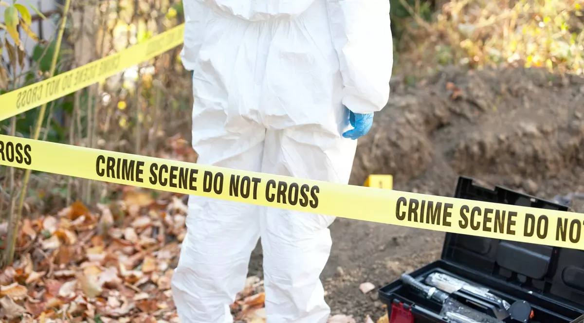 Imagine cu o persoană îmbracată într-un costum alb la locul unei crime. În imagine apare și o bandă galbenă pe care scrie „Locul crimei. Nu treceți”.