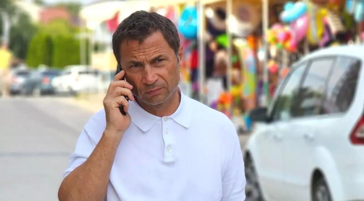 Cristian Radu, îmbrăcat cu un tricou alb, în timp ce vorbește la telefon, pe o stradă, având o mașină albă în spate