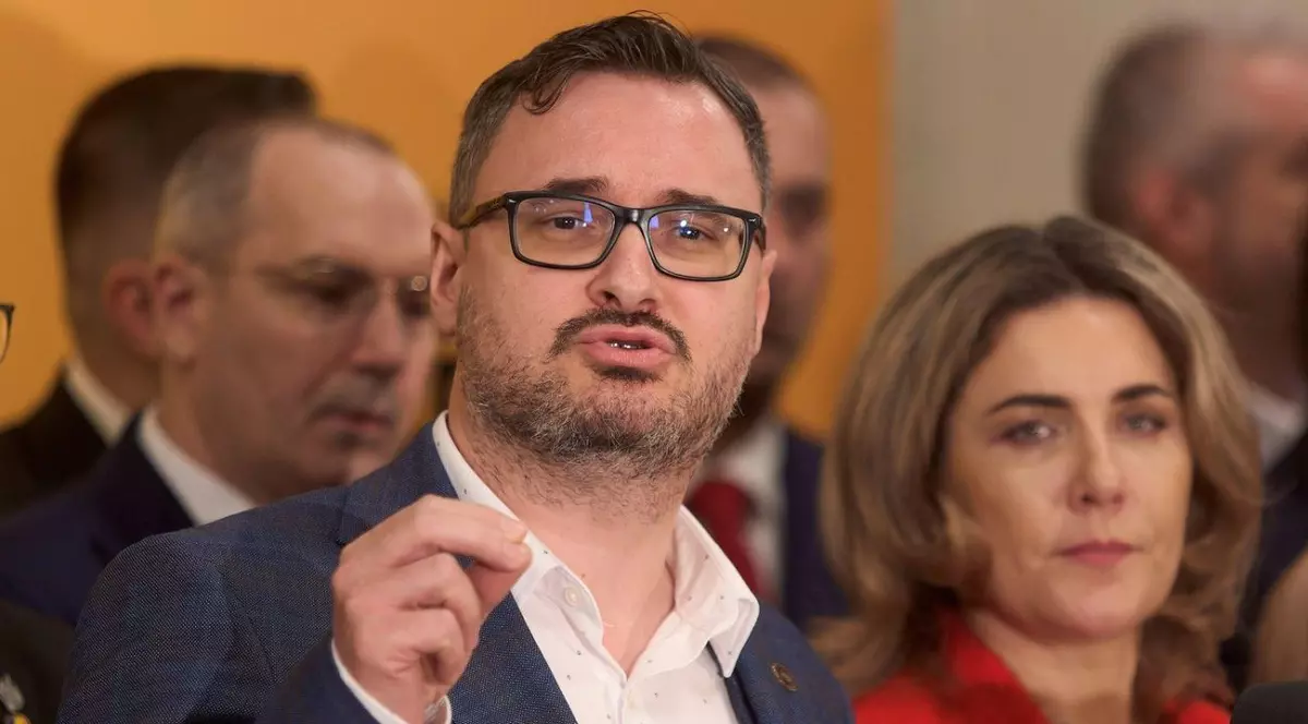 Dan Tanasă, intr-un sacou gri și o cămașă albă, vorbind și gesticulând, ținând o mână ridicată și atingându-și degetul arătător de cel mare