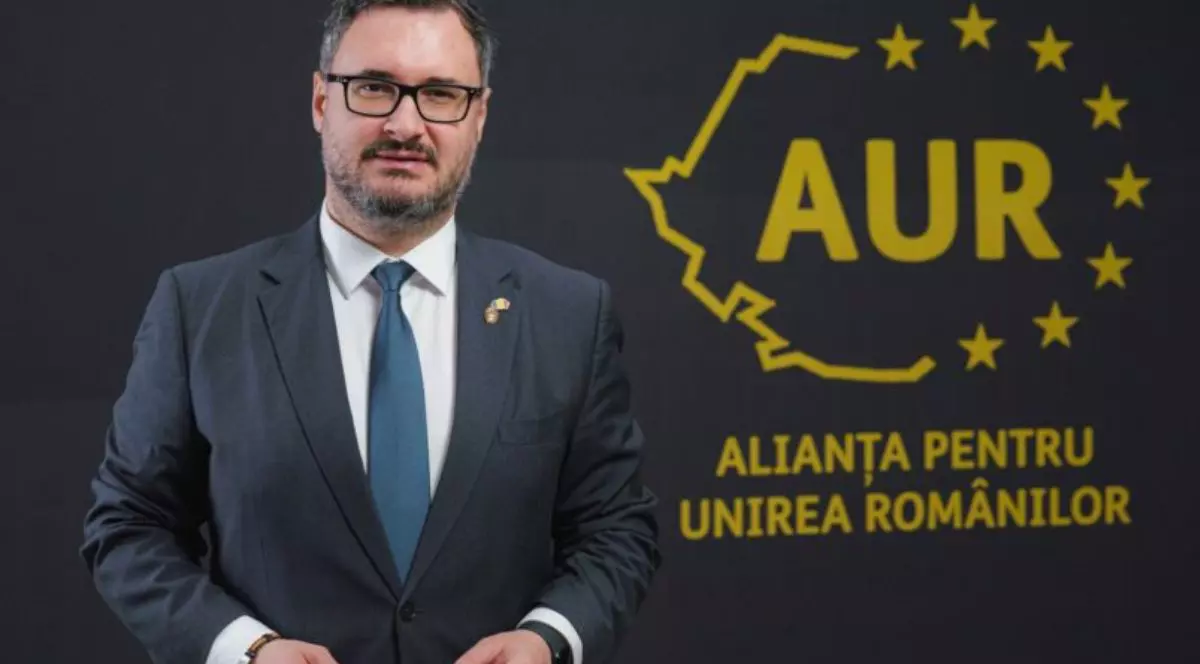 Dan Tanasă, îmbrăcat într-un sacou negru, cămașă albă și o cravată bleumarin, stând cu mâinile pe lângă corp în fața unui fundal negru cu stema aurie a partidului AUR