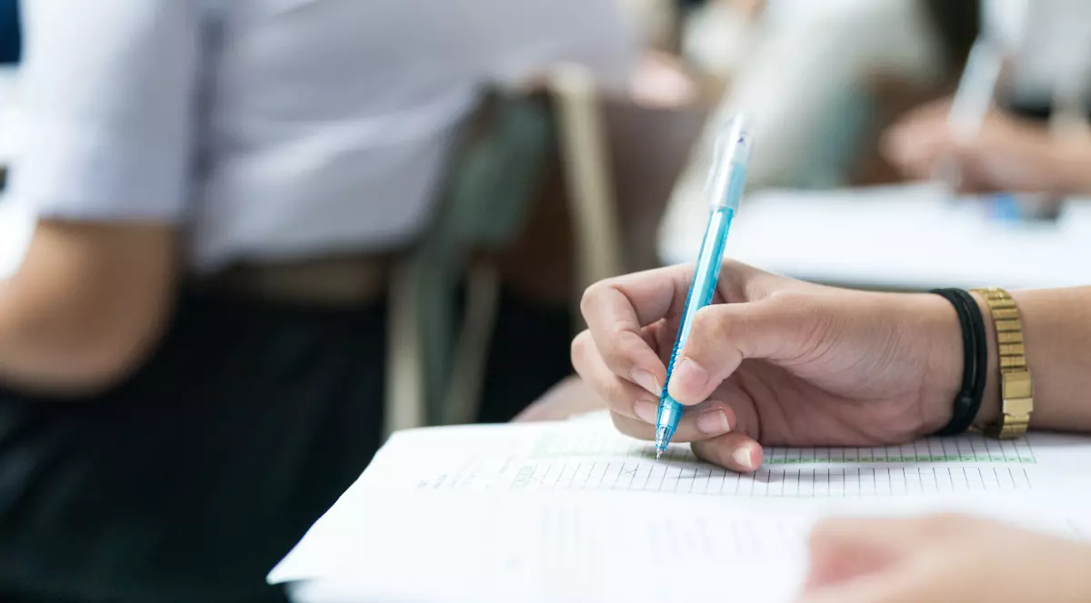 Imaginea arată o persoană care scrie cu un pix albastru pe o foaie, completând un test, într-un cadru ce pare a fi o sală de clasă sau de examen