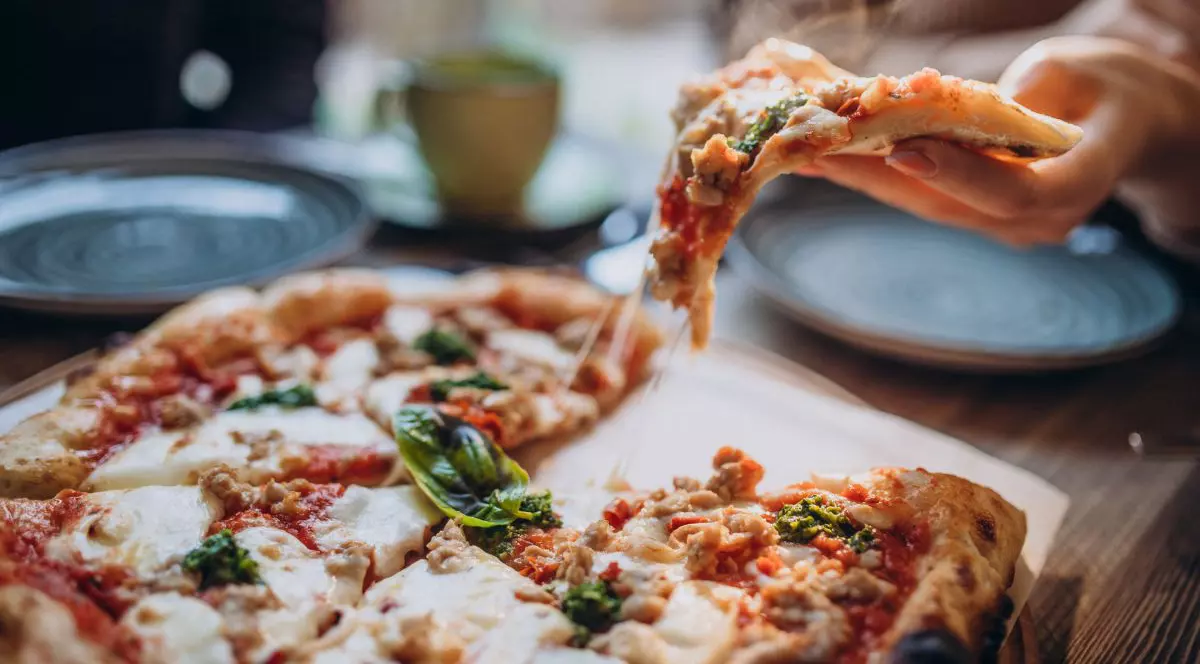 o farfurie cu o pizza tăiată în felii, așezată pe o masă din lemn. O mână ridică o felie de pizza de pe farfurie. Pizza are blat subțire, acoperit cu brânză topită, sos roșu și bucăți mici de ingrediente de culoare maro și verde. Deasupra pizzei, se observă abur. În fundal, se pot vedea două farfurii goale și o cană, toate neclare, cu un decor difuz. Mâna care ține felia de pizza apare în partea dreaptă a imaginii
