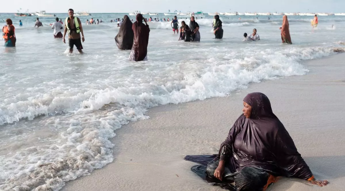 femeie îmbrăcată în port islamic, cu hijab pe cap și haine lungiâ, largi și închise la culoare, stând pe nisip, în fund, la malul mării. în apă se văd alte femei îmbrăcate la fel și un bărbat fără tricou, cu o vestă reflectorizantă