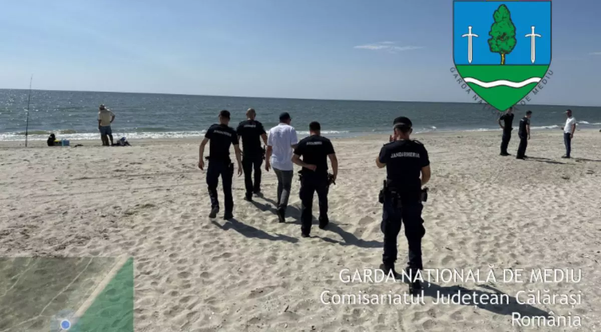 grup de persoane aflate pe o plajă cu nisip. În fața lor este marea cu apă întinsă până la orizont, sub un cer albastru cu câteva nori rari. Cinci bărbați poartă uniforme negre, iar unul dintre ei pare să aibă o șapcă. În stânga, un bărbat stă cu mâinile în șold, iar în plan secund, în partea dreaptă, alți trei bărbați, îmbrăcați diferit, stau și discută. Pe plajă, lângă apă, se află un băț vertical subțire, posibil o undiță