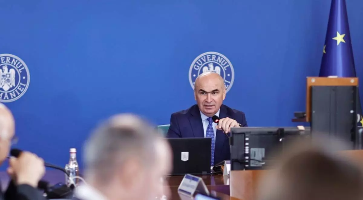 Premierul Ilie Bolojan în timpul unei ședințe de Guvern, acesta vorbește la microfon cu miniștrii prezenți în sală, poartă un costum albastru închis și o cravată albastră cu picățele