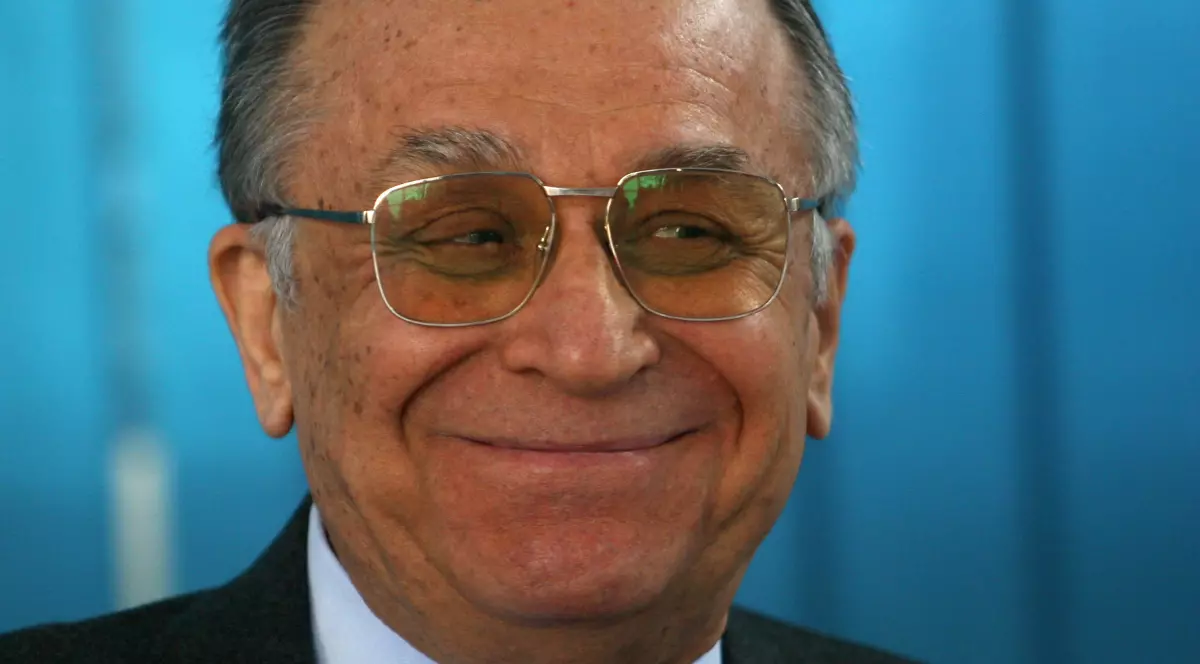 Ion Iliescu, cu ochelari de vedere la ochi, zâmbește, iar în spate are un fundal bleu