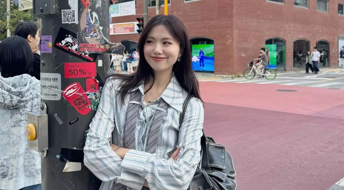 influencerița din Taiwan Irene Gu, purtând o cămașă albă cu dungi gri și o gravată desfăcuta în jurul gâtului, ținând mâinile încrucișate în jurul pieptului și o geantă de piele neagră pe umăr. Femeia stă sprijinită de un stâlp al unui semn de circulație, care este plin de stickere