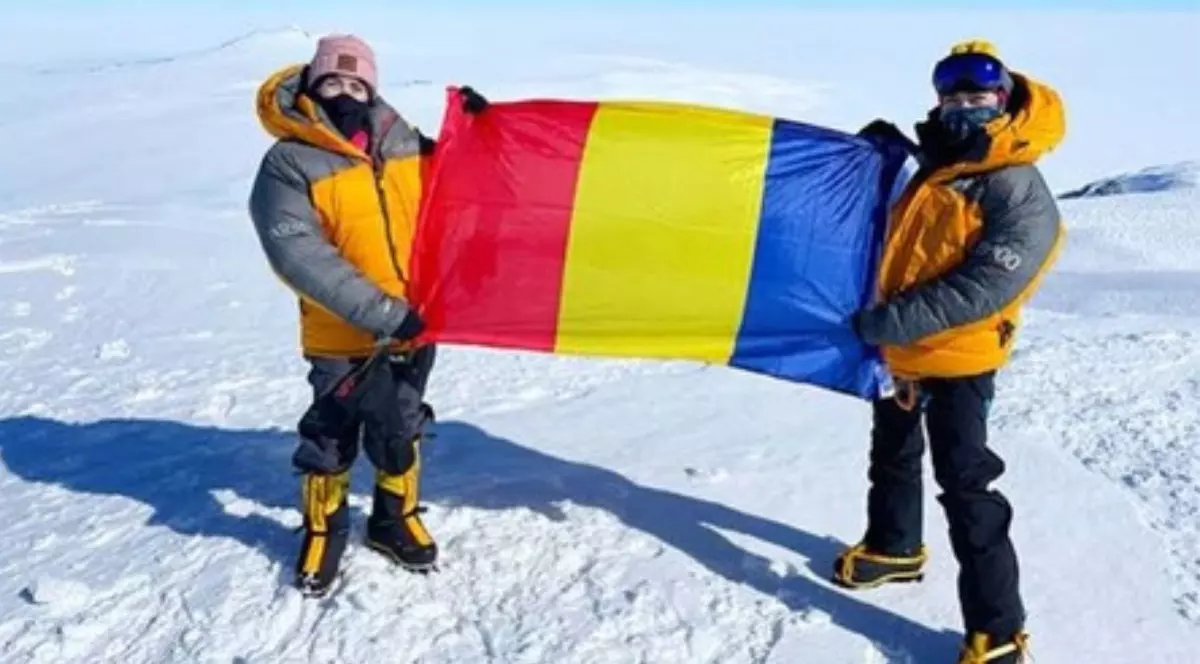 două tinere îmbrăcate în echipament de escaladare montană, format din căciulă, geci portocalii, pantaloni negrii și cizme negre cu dungi portocalii, ținând în mâini drapelul României, pe vârful unui munte