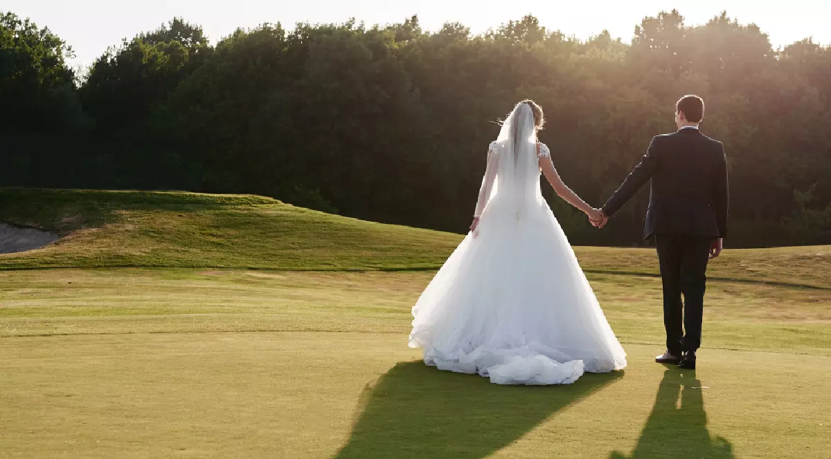 Portret în întregime al mirilor tineri mergând pe iarba verde a terenului de golf la apusul soarelui, vedere din spate.