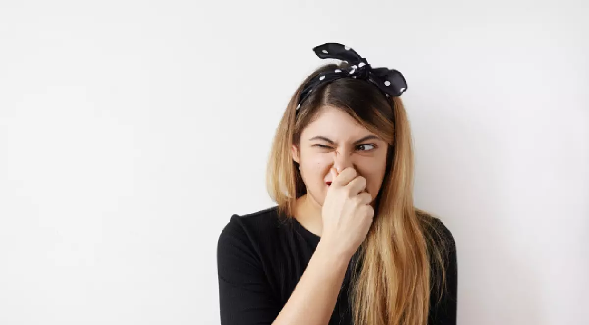 O tânără cu o eșarfă neagră cu buline pe cap, care își ține nasul strâmbându-se, semn că simte un miros neplăcut.