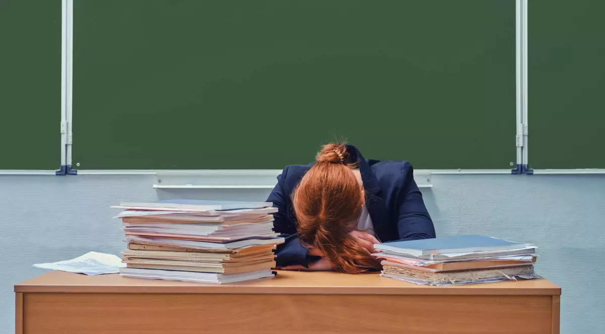 O profesoară care stă cu capul pe catedră, are fața acoperită și părul roșcat, este supărată și înconjurată de cărți