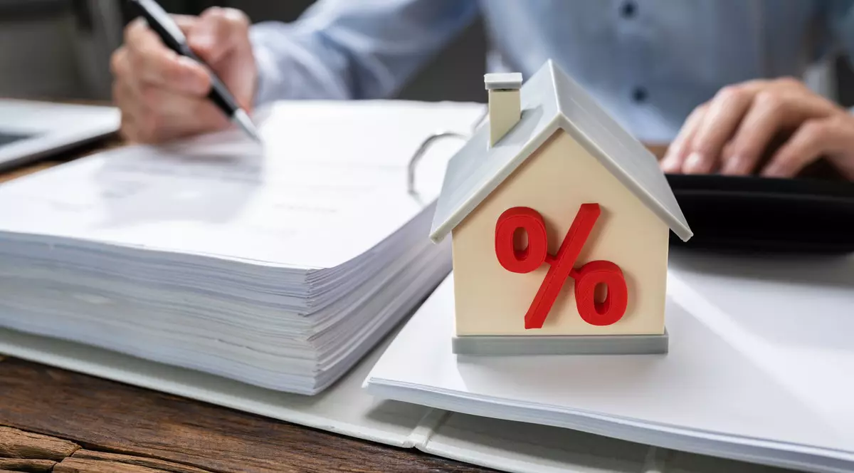 imagine cu caracter ilustrativ. un barbat la birou cu un teanc de hartii in fata si macheta unei case pe care este pus semnul % ca aluzie la creditele imobiliare