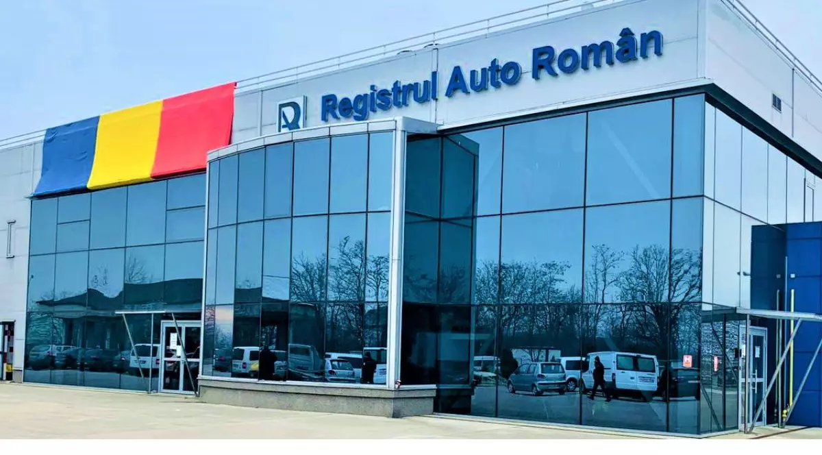 fațada unei clădiri cu ferestre mari, înconjurată de mașini parcate, cu logo-ul și tetul „Registrul Auto Român” scris pe fațadă, alături de steagul României