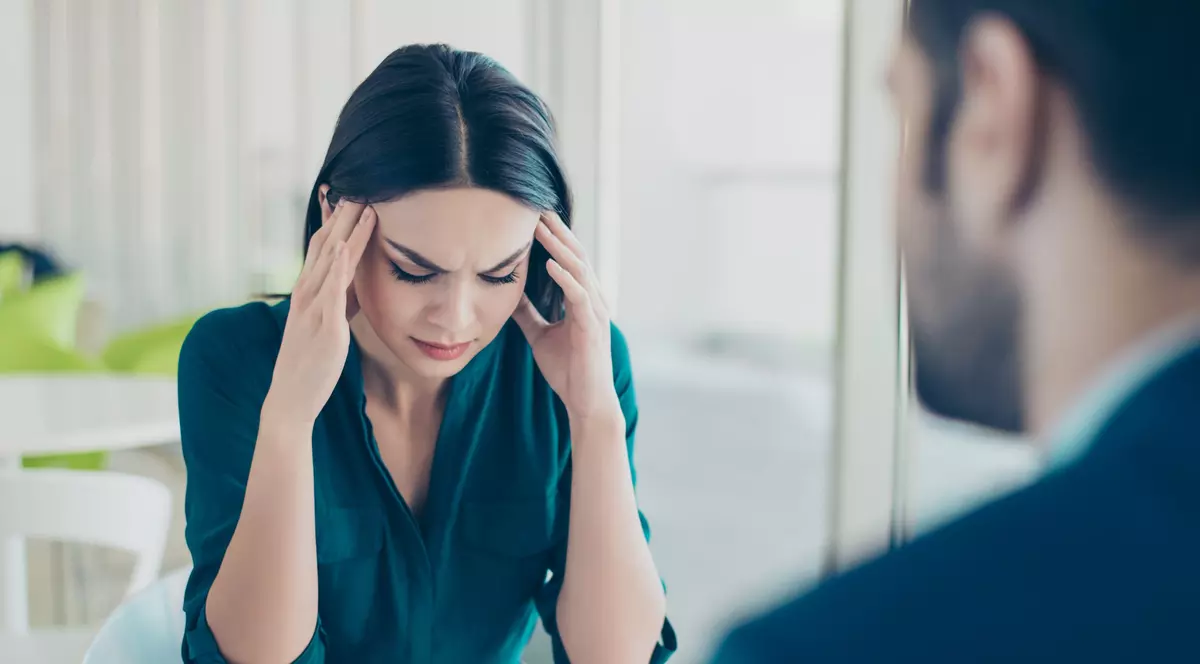 Barbat si femeie care stau fata in fata la masa. Femeia sta cu mainile la tampla, mimand o durere de cap