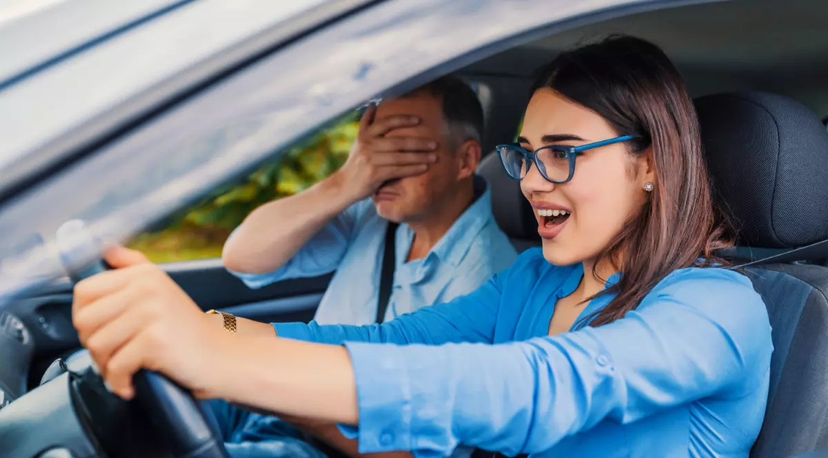 O femeie la volan, în timpul orelor de condus, alături de instructorul auto care își ține mâinile la ochi