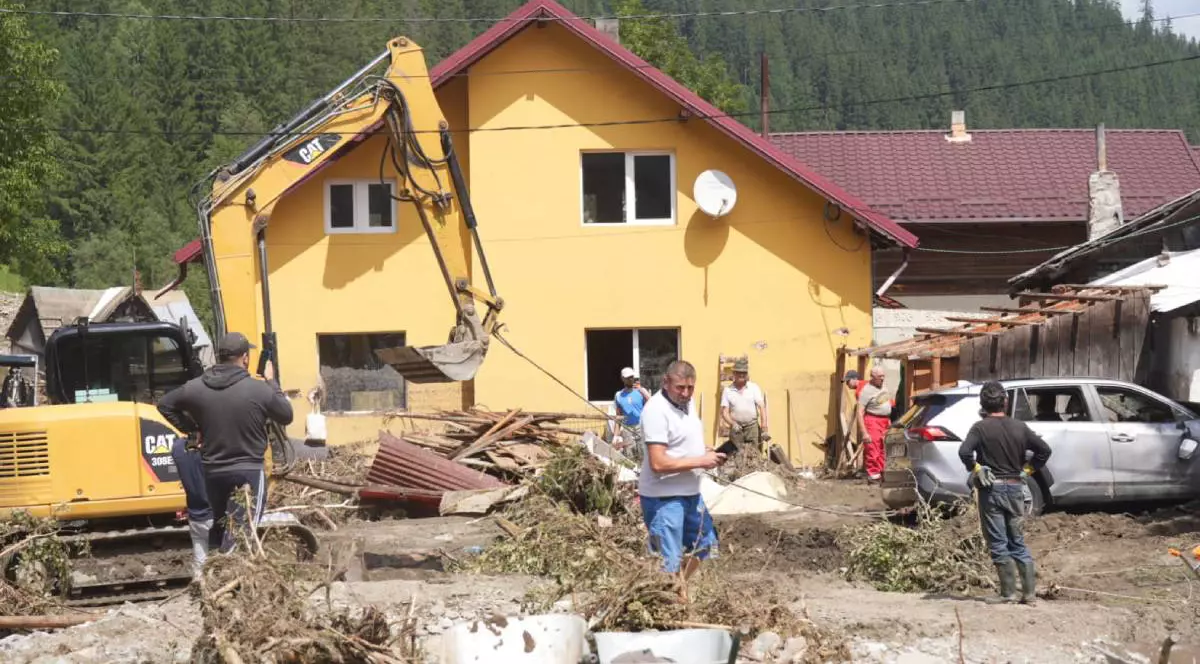 Pompierii și voluntarii de la Broșteni scot nămolul și lemnele din curtea unei case mari, galbene, avariată în timpul inundațiilor