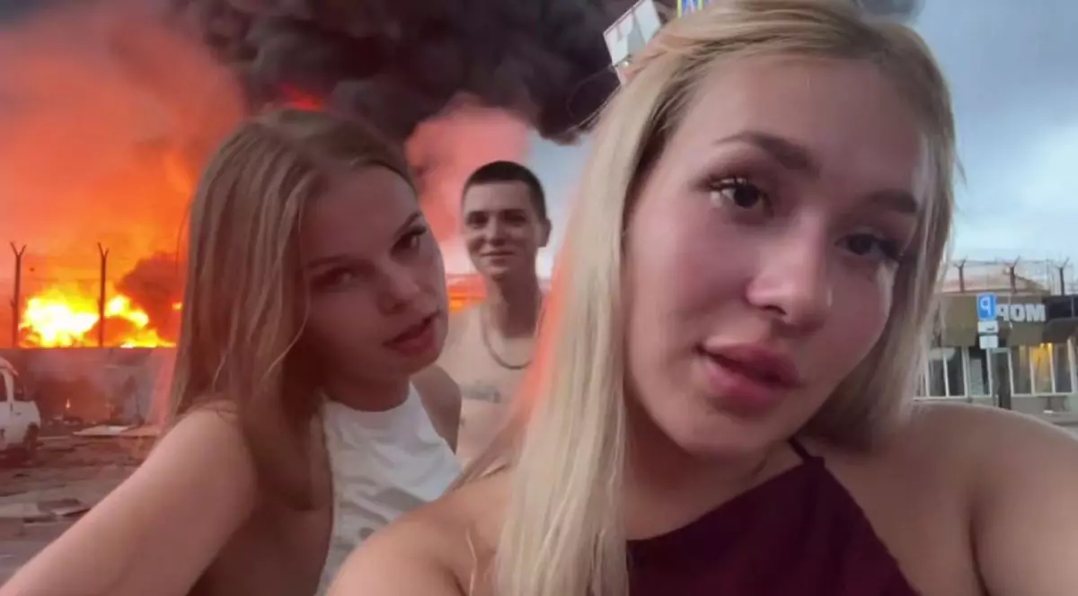 Cei trei tineri din Rusia, în față două fete, una blondă și alta șatenă, iar pe fundal un tânăr și un depozit în flăcări,