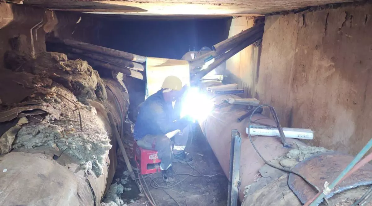 Un muncitor cu cască galbenă pe cap și care stă pe o ladă de bere, într-un subsol, sudează o țeavă