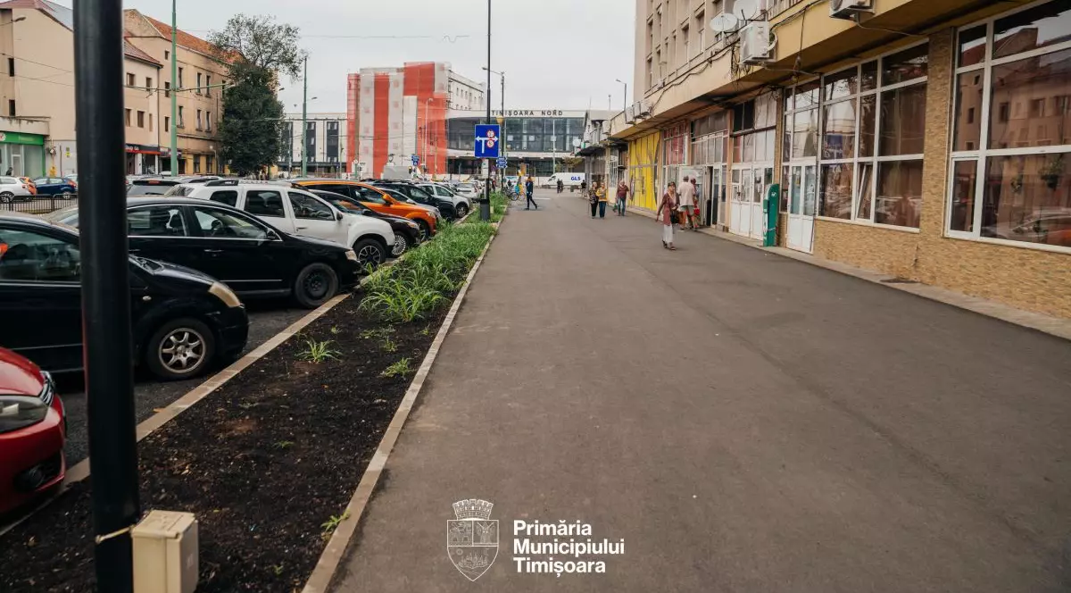 Mai multe pesoane circulă pe un trotur situat lângă un bloc care are magazine și o parcare la parter, în Timișoara, septembrie 2025