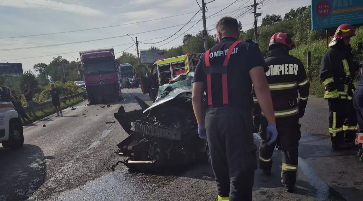 mai mulți pompieri îmbrăcați în uniforme de intervenție, cu veste reflectorizante și căști roșii, care stau pe o șosea largă, în apropierea unei epave de vehicul grav avariat. În fundal se vede un camion de culoare roz în mijlocul drumului, precum și o autospecială cu ușa deschisă. Pe marginea drumului stau câțiva oameni, iar resturi de mașină și fragmente metalice sunt împrăștiate pe carosabil. În lateral se observă și o ambulanță,