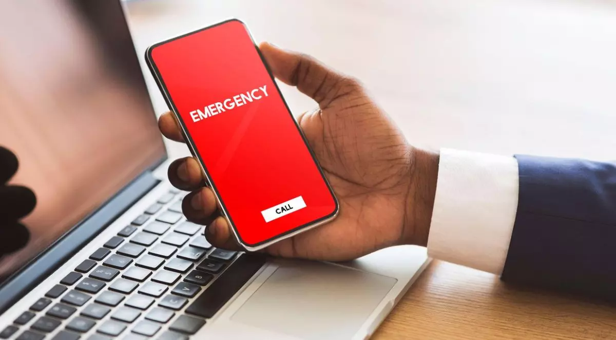 Imagine cu mana unui bărbat cu camasa alba, care tine în mana un telefon cu ecran rosu pe care scrie Emergency, deasupra unui laptop pus pe masă