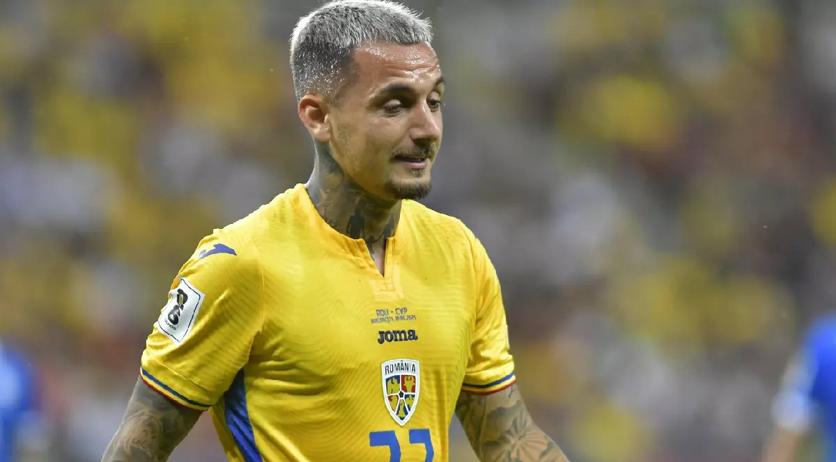 Imaginea este un prim-plan al fotbalistului Alexandru Mitriță. El poartă un tricou de joc al echipei naționale a României, de culoare galbenă, pe care se poate observa emblema Federației Române de Fotbal și un logo JOMA. Are o tunsoare scurtă, cu părul vopsit într-o nuanță deschisă de gri sau blond-cenușiu. O caracteristică vizibilă sunt numeroasele tatuaje care îi acoperă brațele și gâtul. Expresia sa facială este concentrată și serioasă.