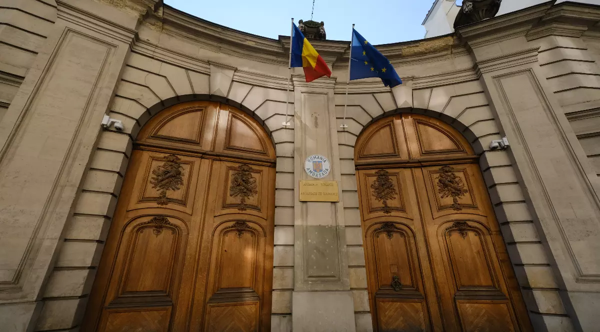 Imaginea arată o clădire cu două uși mari din lemn, identice, fiecare cu un arc rotund în partea superioară. Ușile au sculpturi ornamentale în lemn. Între cele două uși, se află o stemă, probabil a unei instituții. Deasupra intrării, sunt arborate două steaguri: unul este steagul României (roșu, galben și albastru), iar celălalt este steagul Uniunii Europene (albastru cu cerc de stele galbene).