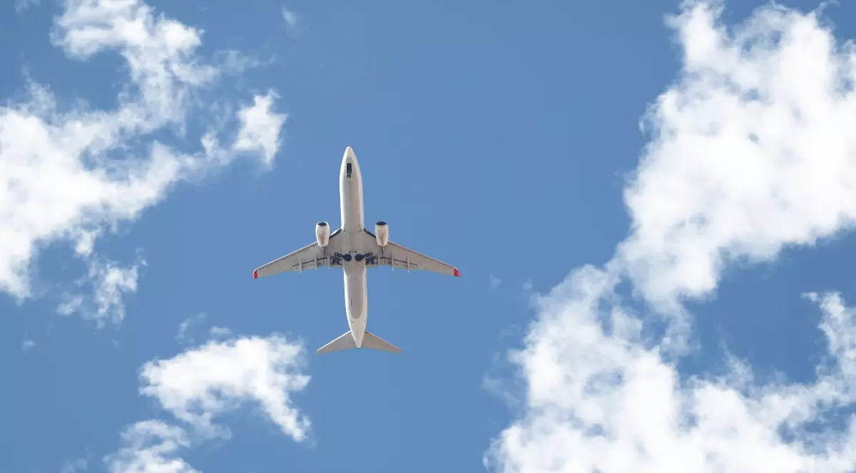 Imaginea arată un avion de pasageri alb, surprins de jos, care zboară pe un cer albastru intens presărat cu nori albi și pufoși. Avionul, care pare să fie la o altitudine joasă, are vizibil trenul de aterizare desfășurat.