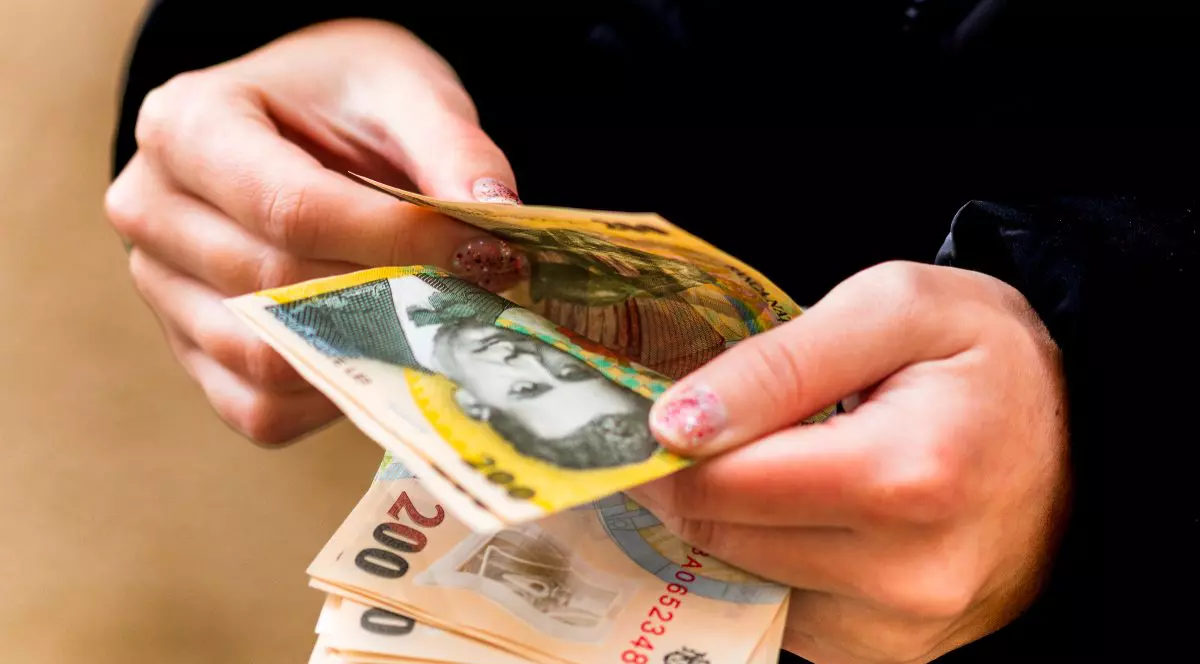 Imaginea, un prim-plan realizat în lumină slabă, surprinde o persoană care numără bancnote românești (Lei), vizibile fiind o bancnotă de 200 de lei și o bancnotă de 10 lei. Mâinile care țin și manevrează banii au o manichiură cu ojă sidefată, pe fundalul unei haine închise la culoare.