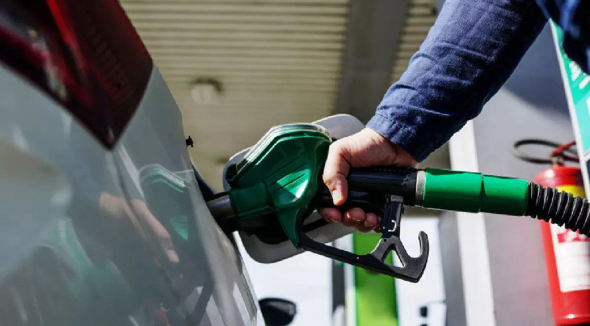 Imaginea, surprinsă de aproape, arată o mână care ține un pistol de alimentare cu combustibil verde și îl introduce în rezervorul unei mașini albe. Fundalul este estompat, dar se pot vedea o pompă de combustibil și un extinctor roșu.